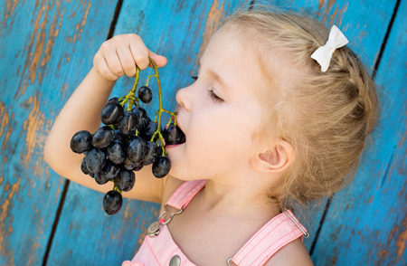 Cute toddler girl eating grapes on a blue wooden backgroundの写真素材