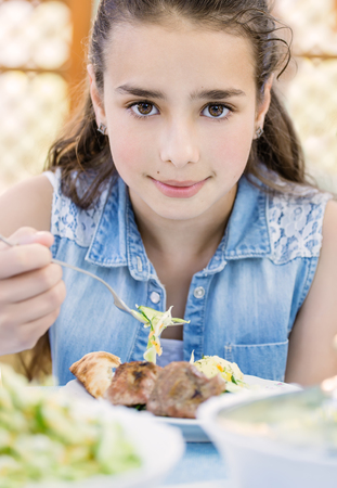 Little beautiful smiling girl with pleasure eats kebab outdoor at the day time. Child having fun near a house. Concept of healthy life.の写真素材