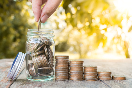 Hand putting coin stack in jar with blurred background and green leaf, Save money and investment conceptの写真素材