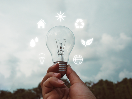Concept save energy efficiency. Hand holding light bulb with icon on blurred tree and sky backgroundの写真素材