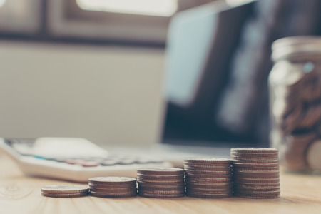 Saving money and investment concept. Money coin stack step staircase growing growth business with jar, laptop and calculator on wood tableの写真素材