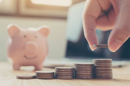 Saving money and investment concept. Hand putting money coin stack step staircase growing growth business with piggy bank, laptop on wood tableの写真素材