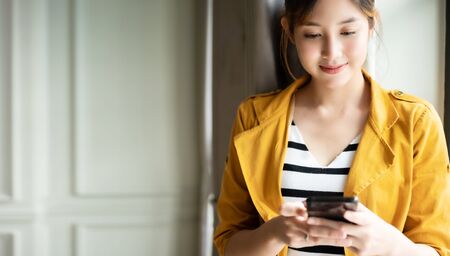Asian woman standing side window with beautiful smile and using application chatting on cell...