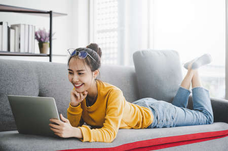 Beautiful young asian woman lying down and using computer laptop talking video conference call on sofa in living roomの写真素材
