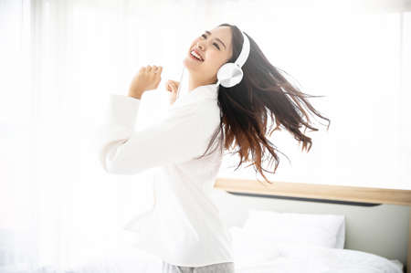Beautiful young asian woman in headphones listening to music and dancing on bed at bedroom in morning timeの写真素材