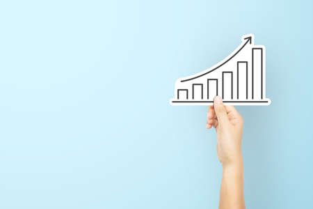 Hand holding paper with arrow and graph increase on light blue background. Business development to success and growing growth conceptの写真素材