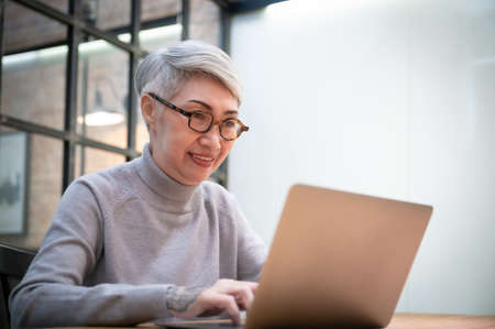 Mature asian business woman wears glasses using laptop computer sit at workplace desk. Happy senior...