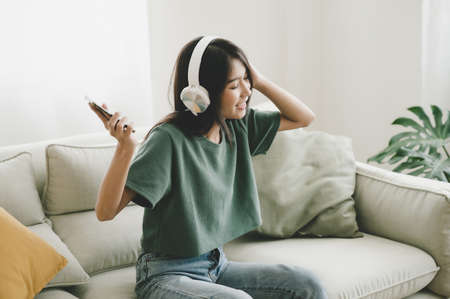 Asian young woman sitting on sofa. She listening music and dancing in living room. She happy and relaxing at free time on weekendの写真素材