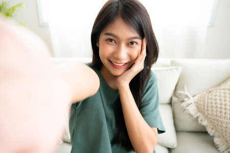Pretty young asian female with big smile sitting at living room. She having fun taking light cheerful selfie on blurred backgroundの写真素材