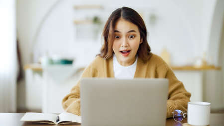 Smiling asian young woman working on laptop at home office. Young asian student using computer remote studying, virtual training, e-learning, watching online education webinar at houseの写真素材