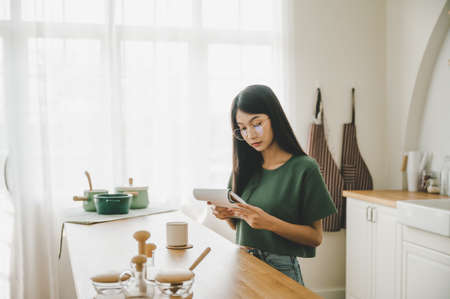 Happy young asian woman wearing glasses in kitchen at home. Female concentrate reading note cooking