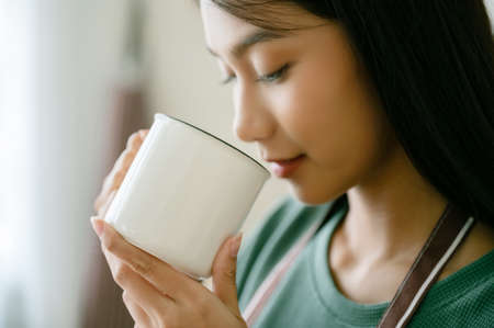 Young asian woman standing beside window and holding mug in kitchen room at home, She drinking milk...