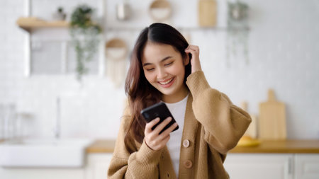 Happy young asian woman relaxing at home. Asia female standing at counter kitchen and using mobile smartphoneの写真素材