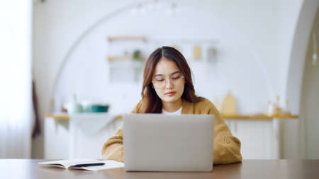 Smiling asian young woman working on laptop at home office. Young asian student using computer remote studying, virtual training, e-learning, watching online education webinar at houseの写真素材