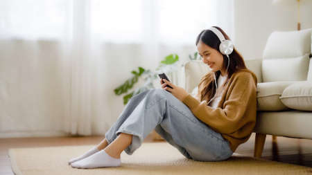 Happy young asian woman relaxing at home. Female smile sitting on sofa and holding mobile smartphone. Girl using video call to friendの写真素材