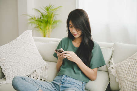Happy young asian woman relaxing at home she is sitting on sofa and using mobile smartphoneの写真素材