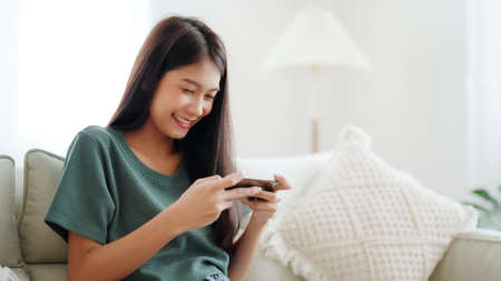 Happy young asian woman relax on comfortable couch at home texting messaging on smartphone, smiling girl use cellphone chatting, browse wireless internet on gadget, shopping online from homeの写真素材