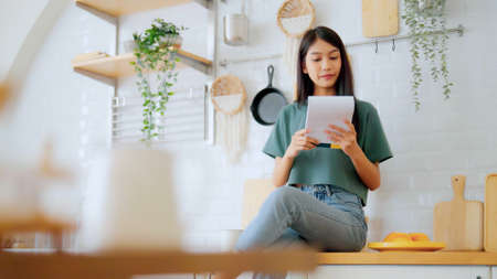Happy young asian woman sitting on counter kitchen at home. Female reading note cookingの写真素材
