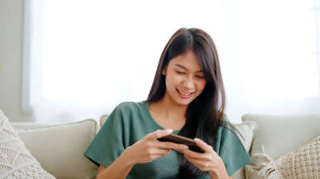 Happy young asian woman relax on comfortable couch at home texting messaging on smartphone, smiling girl use cellphone chatting, browse wireless internet on gadget, shopping online from homeの写真素材