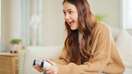 Beautiful young asian woman sitting in living room sofa holding joystick remote control playing online video gameの写真素材
