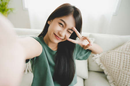Pretty young asian female with big smile sitting at living room. She having fun taking light cheerful selfie on blurred backgroundの写真素材