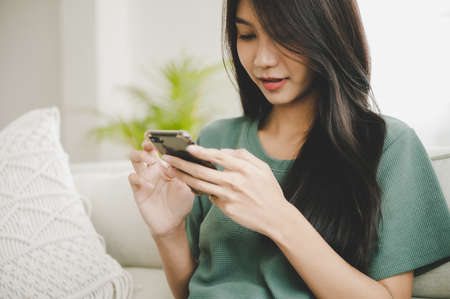 Happy young asian woman relaxing at home she is sitting on sofa and using mobile smartphoneの写真素材