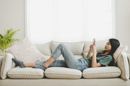 Happy young asian woman relaxing at home she is lying down on sofa and using mobile smartphoneの写真素材