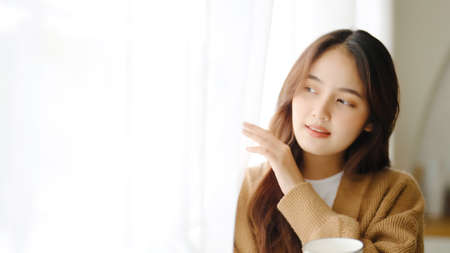 Young asian woman standing beside window and holding mug in living room at home, She drinking milk after wake up in the morningの写真素材