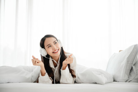 Beautiful young asian woman in headphones listening to music and lying down on bed at bedroom in morning timeの写真素材
