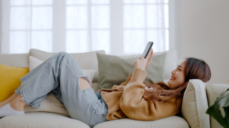 Happy young asian woman relax on comfortable couch at home texting messaging on smartphone, smiling girl use cellphone, browse wireless internet on gadget, shopping online from homeの写真素材