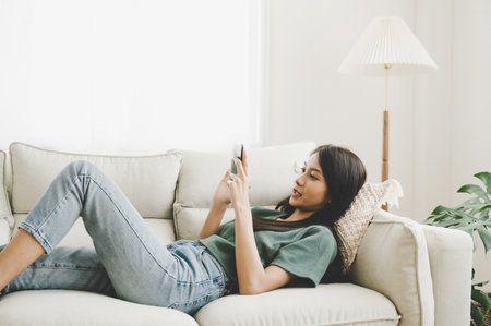 Happy young asian woman relaxing at home. Female is lying down on sofa and using mobile smartphoneの写真素材