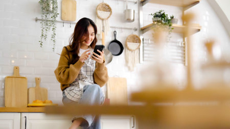 Pretty young asian female smile and sitting on counter kitchen room. She using mobile smart phone having fun video call talking with her friendの写真素材