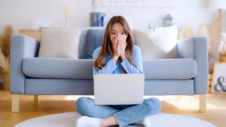 Young asian woman in good spirits working on laptop at home while sitting on the floor close to the couch. Excited female winner celebrating success while glancing at notebookの写真素材