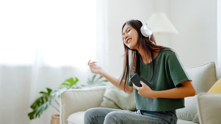 Young asian woman listening to music on couch in living room at home. Happy asia female using mobile smartphone, wearing headset and sitting on sofaの写真素材