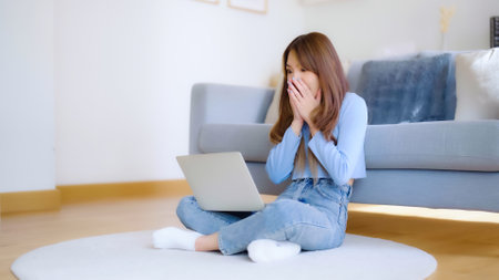 Young asian woman in good spirits working on laptop at home while sitting on the floor close to the couch. Excited female winner celebrating success while glancing at notebookの写真素材