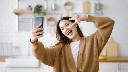 Pretty young asian female smile and stand at kitchen room. She having fun taking selfie and video call talking with her friendの写真素材