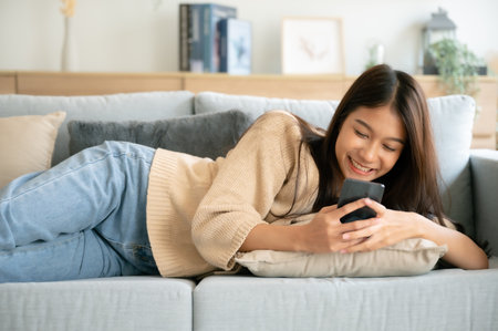 Happy young asian woman relax on comfortable couch at home texting messaging on smartphone, smiling girl use cellphone, browse wireless internet on gadget, shopping online from homeの写真素材