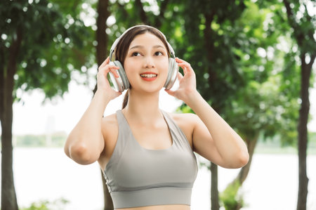 Asian woman trainer is engaged in fitness in public park. Female wearing sport bra and using headphone listening to music. Healthy lifestyle, sports outdoor activities in parkの写真素材