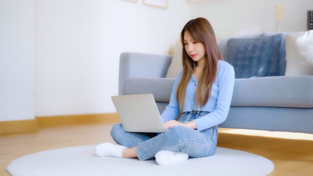 Young asian woman in good spirits working on laptop at home while sitting on the floor close to the couchの写真素材
