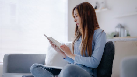 Young asian woman reading a book while seated on couch at homeの写真素材