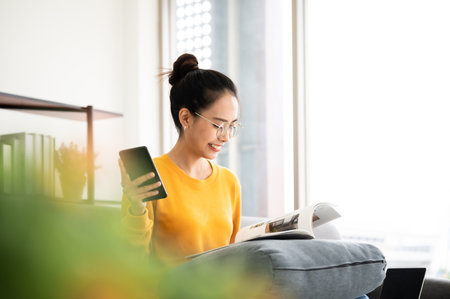 Happy young asian woman reading on the sofa and holding mobile smartphone in living room at homeの写真素材
