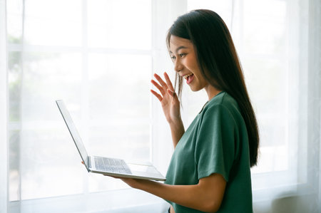 Happy smiling asian young woman using work laptop and standing beside window at homeの写真素材