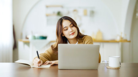 Young asian woman sitting at table and taking notes in notebook. Working on table with computer laptop, document paperwork and cup of coffee. Student learning onlineの写真素材