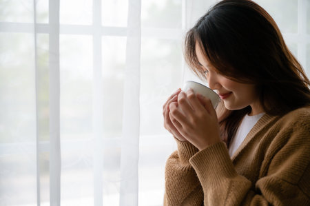 Young asian woman standing beside window and holding mug in living room at home, She drinking milk after wake up in the morningの写真素材