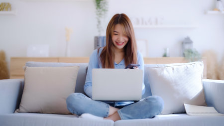 Young asian woman using computer laptop and mobile smartphone while seated on couch at homeの写真素材