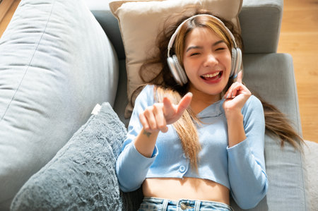 Young asian woman listening to music on couch in living room at home. Happy asia female using mobile smartphone, wearing headset and lying down on sofaの写真素材