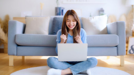Young asian woman in good spirits working on laptop at home while sitting on the floor close to the couch. Excited female winner celebrating success while glancing at notebookの写真素材