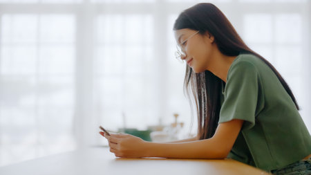 Happy young asian woman wearing glasses and relaxing at home. Asia female standing at counter kitchen and using mobile smartphoneの写真素材