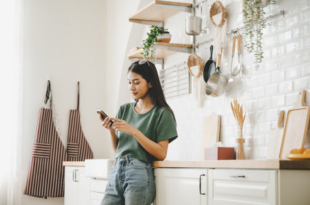 Happy young asian woman relaxing at home she is standing at counter kitchen and using mobile smartphoneの写真素材