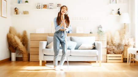 House cleaning with fun. Happy young asian housewife singing her favorite song during cleanup, using mop as microphone, enjoying domestic work. Young woman dancing and cleaning in living roomの写真素材